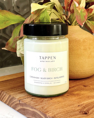 Candle jar labeled 'Fog & Birch' by Tappen Apothecary on a wooden surface with leaves in the background.