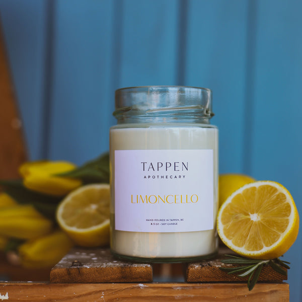 Candle jar labeled 'Tappen Apothecary Limoncello' with lemons on a wooden surface against a blue background