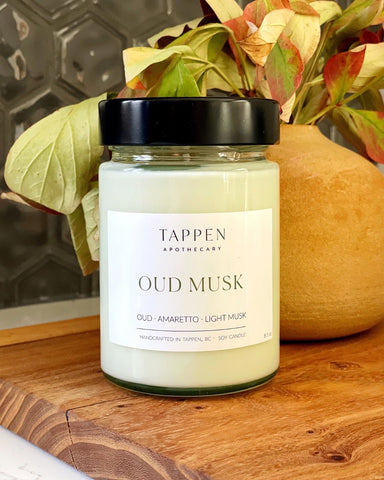 Tappen Apothecary Oud Musk candle on a wooden surface with decorative plants in the background