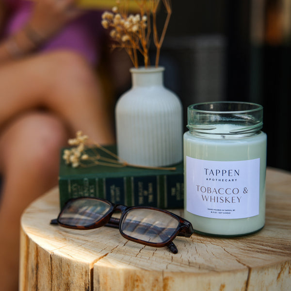 Candle labeled 'Tappen Tobacco & Whiskey' on a wooden surface with sunglasses and a vase in the background.